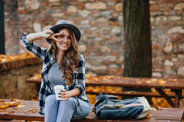 Lovable white female model with curly hairstyle posing with peace sigh while drinks coffee in park