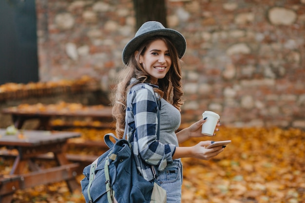 Lovable white girl in checkered shirt holding smartphone and cup of coffee in september park