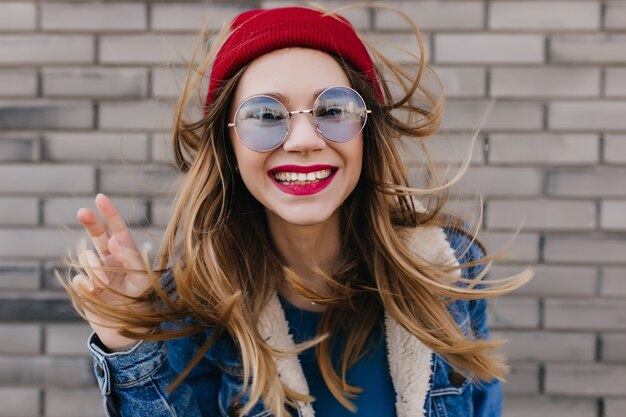 Lovable woman in casual blue glasses fooling around during outdoor photoshoot. Portrait of excited blonde girl with bright makeup posing on brick wall.