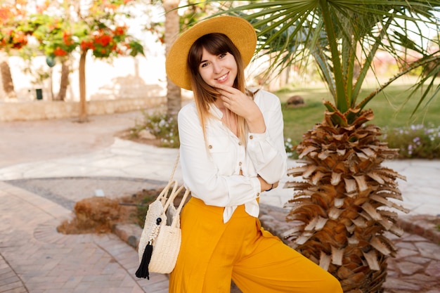 Бесплатное фото Привлекательная женщина в соломенной шляпе и белой блузке позирует в тропическом отеле во время отпуска.