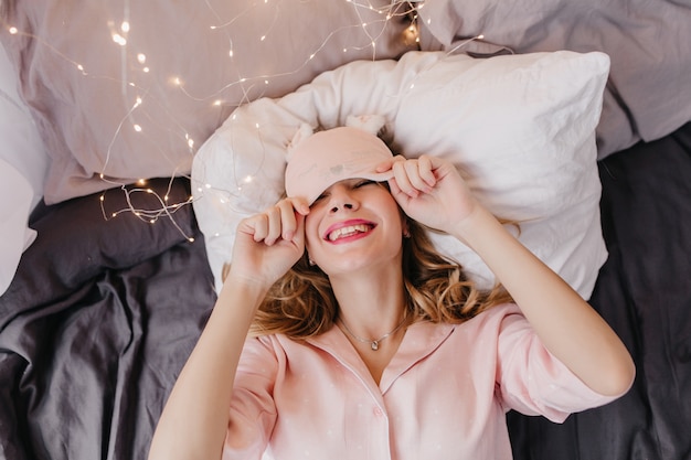 Очаровательная молодая женщина в розовом ночном костюме позирует в постели с улыбкой. Возбужденная кавказская девушка в маске для глаз расслабляется утром и смеется.