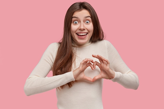 Love and care concept. Beautiful woman smiles broadly, shows heart sign over chest, wears casual clothes, has happy exxpression, stands against pink background