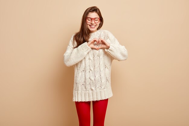 Love concept. Happy smiling girlfriend makes heart gesture with fingers, expresses good feelings, dressed in white jumper, isolated over beige wall. Sign language concept. Symbol of charity