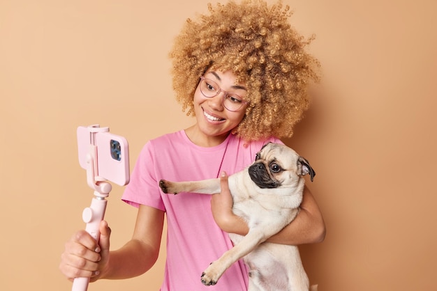 Love friendship between pet and owner positive curly blonde woman poses for selfie with pug dog use smartphone on stick make photo isolated over beige background girl has new member of family