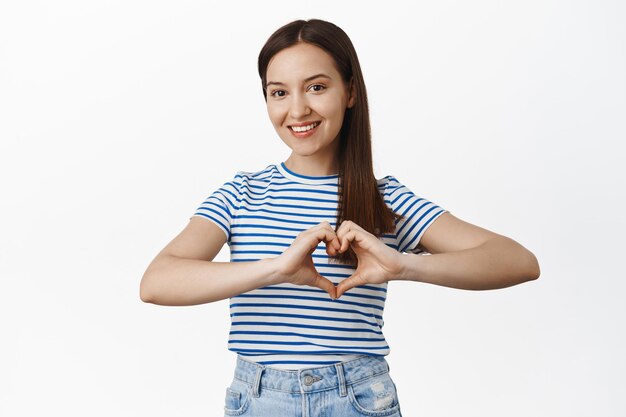 Love is around. Smiling beautiful girl shows heart gesture near chest, express like or sympathy, passionate about smth, standing against white background in t-shirt