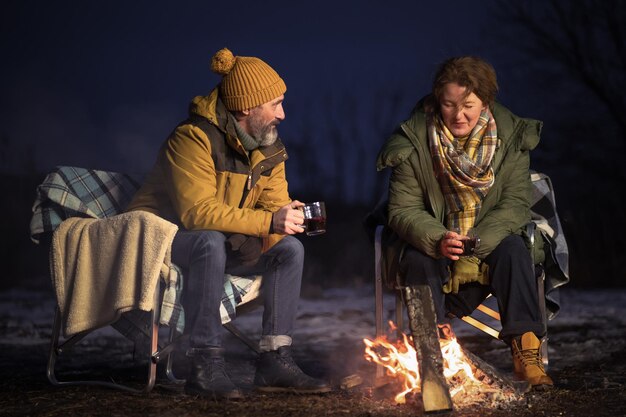 冬の旅で格子縞の毛布の上に座って熱い自然のお茶を飲むキャンプファイヤーで成熟したカップルのロマンチックなディナーを愛して雪の中でキャンプファイヤーに座っているカップル家族での休暇と旅のコンセプト