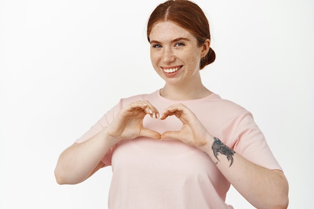 In love. young smiling redhead female shows heart sign and looks happy, like something, express affection or sympathy, standing in tshirt on white