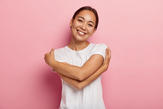 Free photo love yourself. pretty glad asian girl embraces herself, feels comfort and care, tilts head, wears white t shirt, has no make up, isolated over rosy wall, thinks about lover, wants be in his warm arms