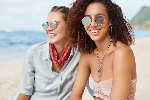 어두운 피부와 곱슬 머리를 가진 사랑스러운 아프리카 계 미국인 여성은 긍정적 인 미소를 가지고 있으며 선글라스를 착용하고 바다 경치를 즐기는 여자 친구 근처에 앉아 해변에서 차분한 분위기를 즐깁니다. 우정 개념