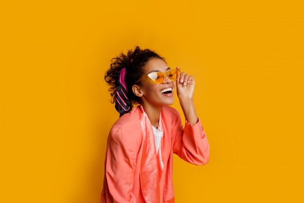 Симпатичная африканская девушка в розовой куртке позирует с счастливым выражением лица на желтом фоне.