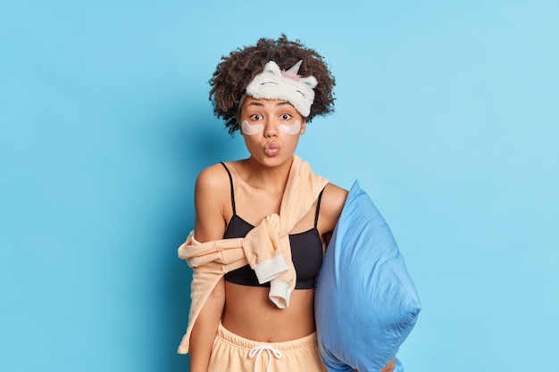 Lovely Afro American millennial girl keeps lips rounded as wants to kiss you says good night wishes sweet dreams dressed in pajama isolated over blue wall