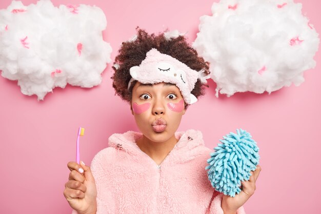 lovely afro american woman keeps lips folded wants to kiss you holds toothbrush and bath sponge dressed in nightwear poses indoor