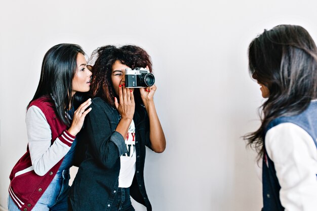 Симпатичная азиатская девушка выглядит как очаровательный африканский фотограф фотографирует свою подругу. Брюнетка молодая женщина позирует для камеры перед фигурной дамой в черном наряде.