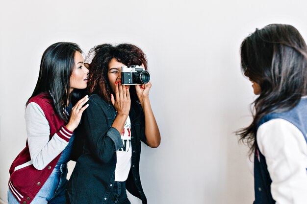 Lovely asian girl looks how charming african photographer taking photo of her friend. Brunette young woman posing for camera in front of curly lady in black outfit.