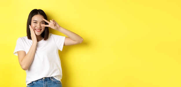 Lovely asian girl in white tshirt posing with hand on cheek showing peace sign on eye standing over