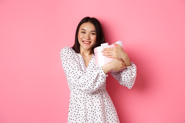 Lovely asian woman hugging her gift and smiling thankful receive valentines day present standing ove...