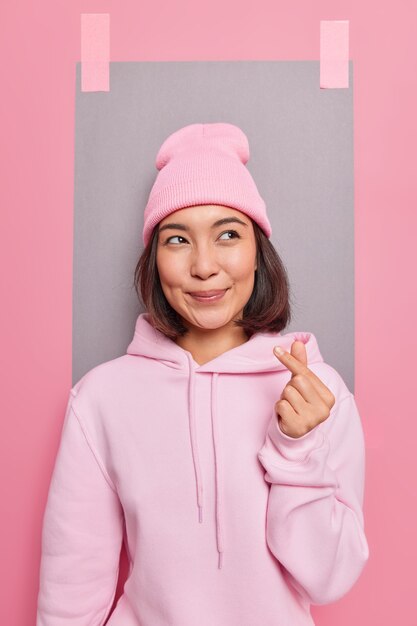 Lovely Asian woman makes korean like sign demonstrates truthful feelings and love shows finger heart gesture dressed in casual hoodie and hat poses against studio wall with plastered sheet of paper