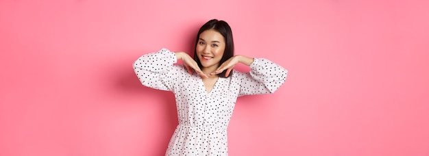 Free photo lovely asian woman showing her beautiful clean face smiling pretty standing in dress against pink ro