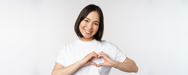 사랑스러운 아시아 여성이 배경 위에 서 있는 부드러움과 애정을 표현하는 하트 사인을 보여주며 웃고 있습니다.