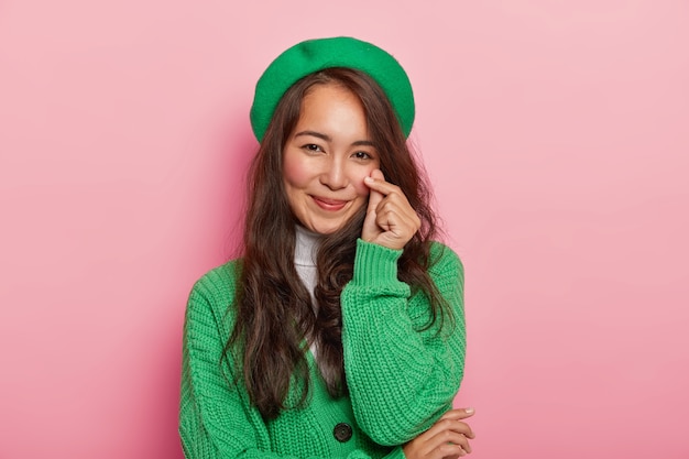 Lovely attractive brunette woman makes korean like gesture, shapes little heart with fingers, has long dark straight hair, wears green beret and jumper on buttons