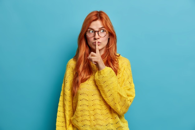 Lovely attractive redhead woman makes silence gesture keeps index finger to lips makes hush sign wears optical glasses and yellow sweater.