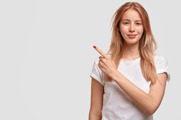 Lovely beautiful woman with satisfied expression, has straight hair, points aside at upper left corner with fore finger, dressed in casual domestic clothes, stands against white wall.