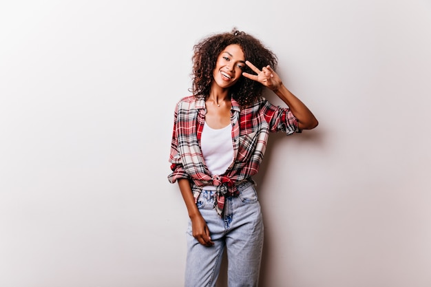 Bella ragazza nera in pantaloni denim vintage in posa con il segno di pace. modello femminile entusiasta in camicia a scacchi isolato su bianco.