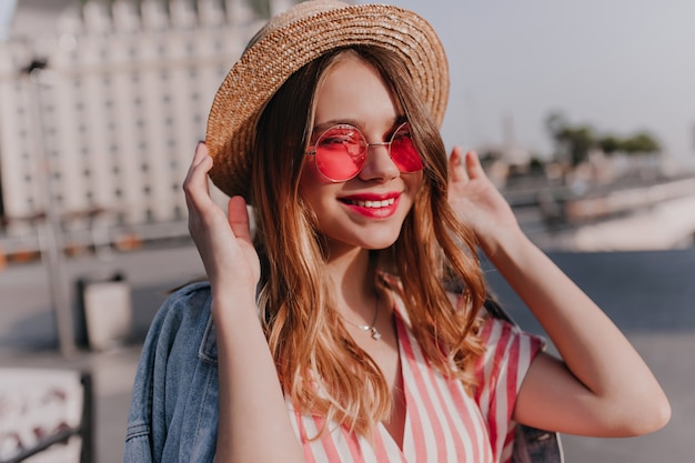 Lovely blithesome girl in pink glasses posing with gently smile. Outdoor shot of stylish white woman touching her straw hat on city.