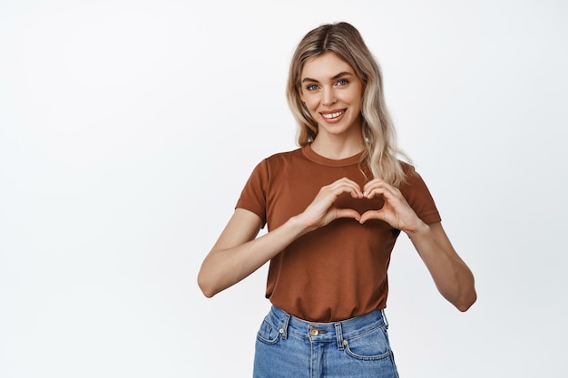 Lovely blond girl shows heart sign near chest and smiling standing flirty against white background