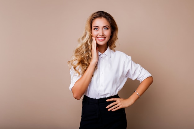 Lovely blond woman with perfect smile in white blouse posing over beige wall. Stylish workwear outfit.