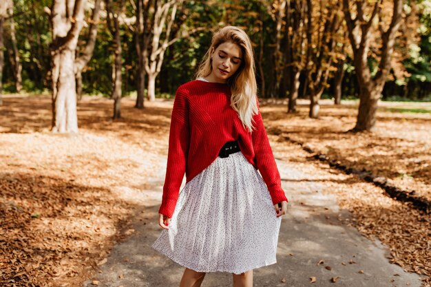 Lovely blonde girl posing in the autumn park. Wearing beautiful white dress with nice red sweater.