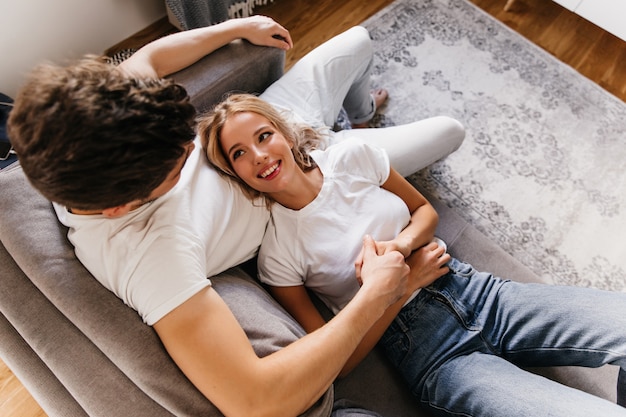 Lovely blonde girl with dreamy look holding hand of her man. Sweet couple on sofa spending time with each other.