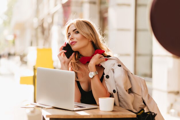 Lovely blonde girl in wristwatch talking on phone while working with laptop in cafe