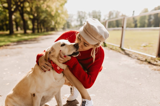 Милая блондинка нежно целует собаку на дорожке в осеннем парке. Стильная девушка хорошо себя чувствует под солнцем.