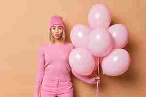 Free photo lovely blonde woman keeps lips rouded sends airkiss to camera comes on birthday party holds bunch of inflated helium balloons wears pink clothes isolated over brown background celebration concept