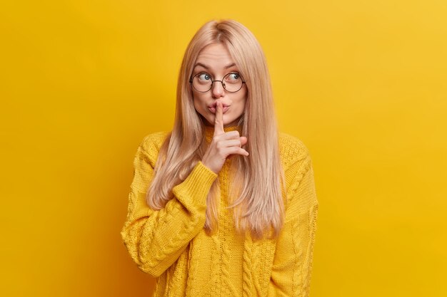 Lovely blonde woman makes silence gesture hides secret tells confidential information has mysterious expression dressed in casual wear asks not to make noise makes shh sign. Shushing and secrecy