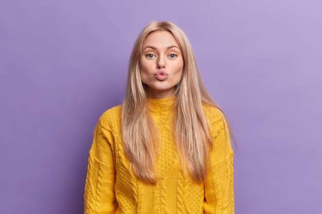 Lovely blonde young woman folded lips blow kiss has romantic face expression expresses sympathy confesses in love to boyfriend wears casual yellow sweater 
