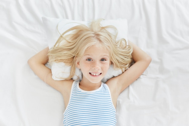 Foto gratuita adorabile bambina dagli occhi blu con le lentiggini, sdraiata sul cuscino bianco, tenendosi le mani dietro, sorridendo piacevolmente, essendo felice di vedere i suoi genitori nella sua camera da letto. bambina che riposa nel letto