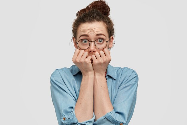 Lovely blue eyed female feels scared, bites fingernails with fear, feels insecure, wears spectacles and denim jacket, isolated over white wall. People and anxiety concept