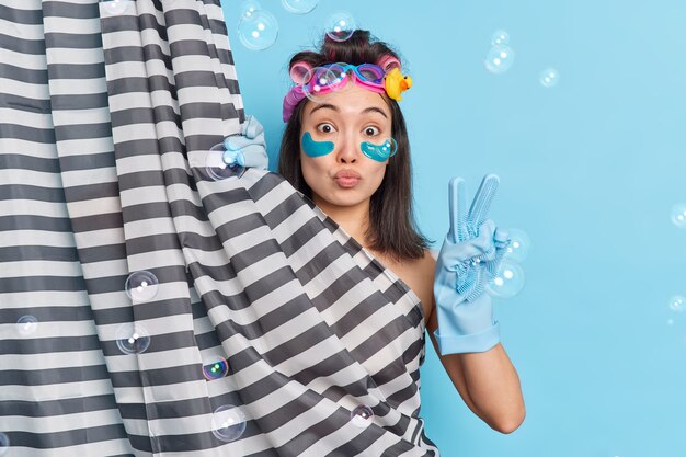 Lovely brunette Asian woman applies collagen patches while taking douche makes peace gesture undergoes beauty procedures hides behind shower curtain poses against blue wall soap bubbles around