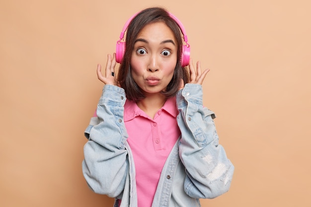 La bella donna asiatica bruna con i capelli scuri tiene le labbra piegate ha un'espressione scioccata ascolta musica tramite cuffie wireless rosa indossa una giacca di jeans isolata sul muro beige