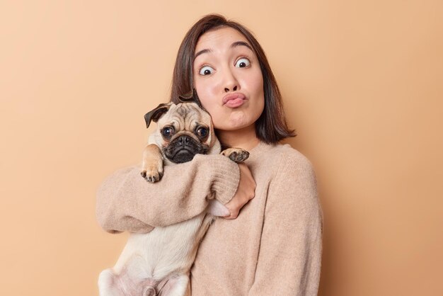 Lovely brunette young Asian woman makes funny grimace keeps lips rounded embraces pug dog with love going to have walk togehter enjoys company of pet wears jumper isolated over beige background.