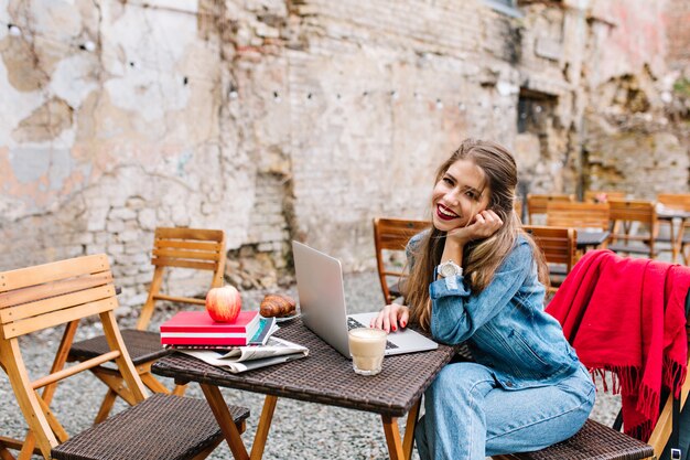 Прекрасная деловая женщина с длинными светлыми волосами, используя белый портативный компьютер на обеденном перерыве в открытом кафе на фоне кирпичной стены. Красивая девушка в джинсах, сидя за деревянным столом.