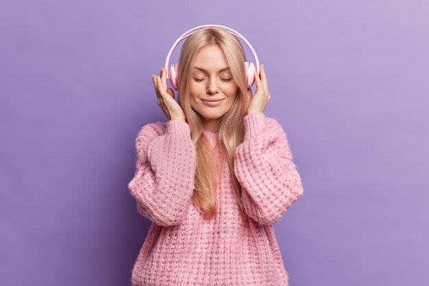 Lovely calm blonde woman keeps hands on stereo headphones keeps eyes closed listens music enjoys every tune dressed in knitted jumper 