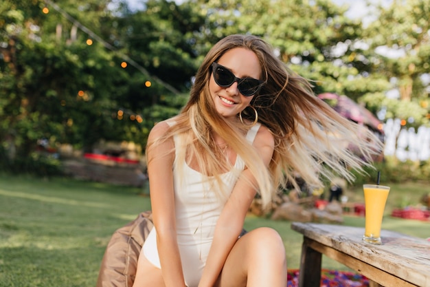 Free photo lovely carefree woman with long hair posing on nature and smiling. romantic tanned female model sitting in park with glass of cold orange juice.