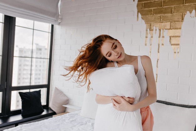 Lovely caucasian female model posing with pillow. Indoor photo of graceful ginger woman having fun in morning.