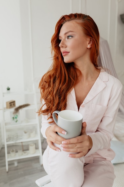朝のコーヒーを保持しているピンクのパジャマを着ている赤い髪の女性と素敵な白人の女の子