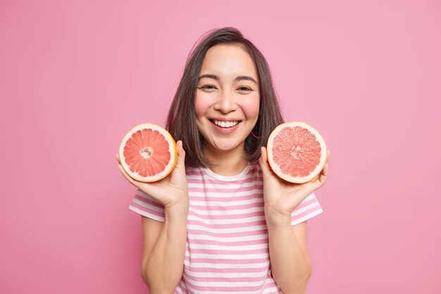 Милая жизнерадостная азиатская женщина с темными волосами держит два ломтика грейпфрута, использует цитрусовые для изготовления натурального косметического продукта, небрежно одетая, имеет счастливое выражение на розовой стене.