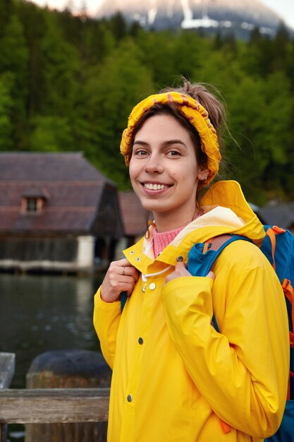 Lovely cheerful lady with pleasant smile, wears yellow raincoat with hood, carries rucksack