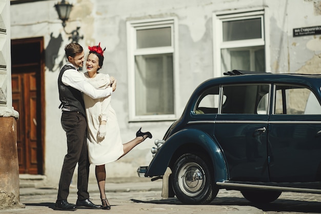 Free photo lovely couple dressed in 30's style hugs on the street standing before an old car